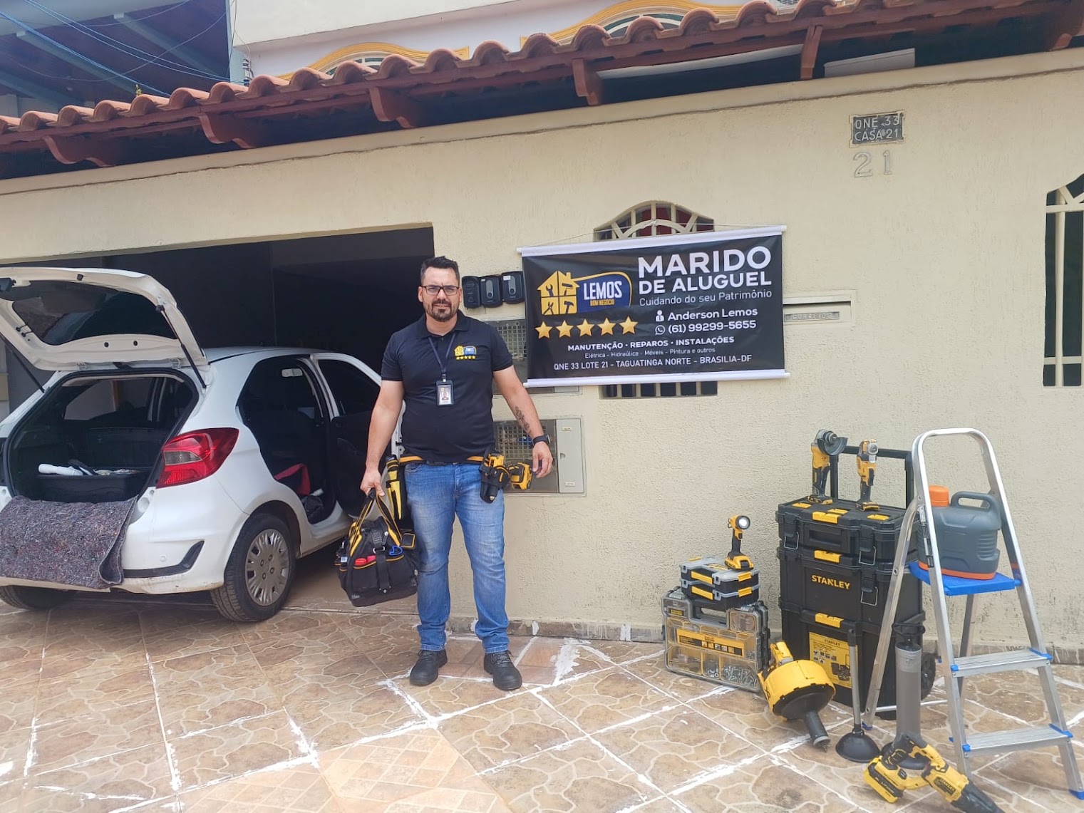 Anderson Lemos com maleta de ferramentas, caixas de ferramentas e escada ao lado do veículo de atendimento, em frente ao banner da Lemos Bom Negócio em Taguatinga Norte, Brasília-DF